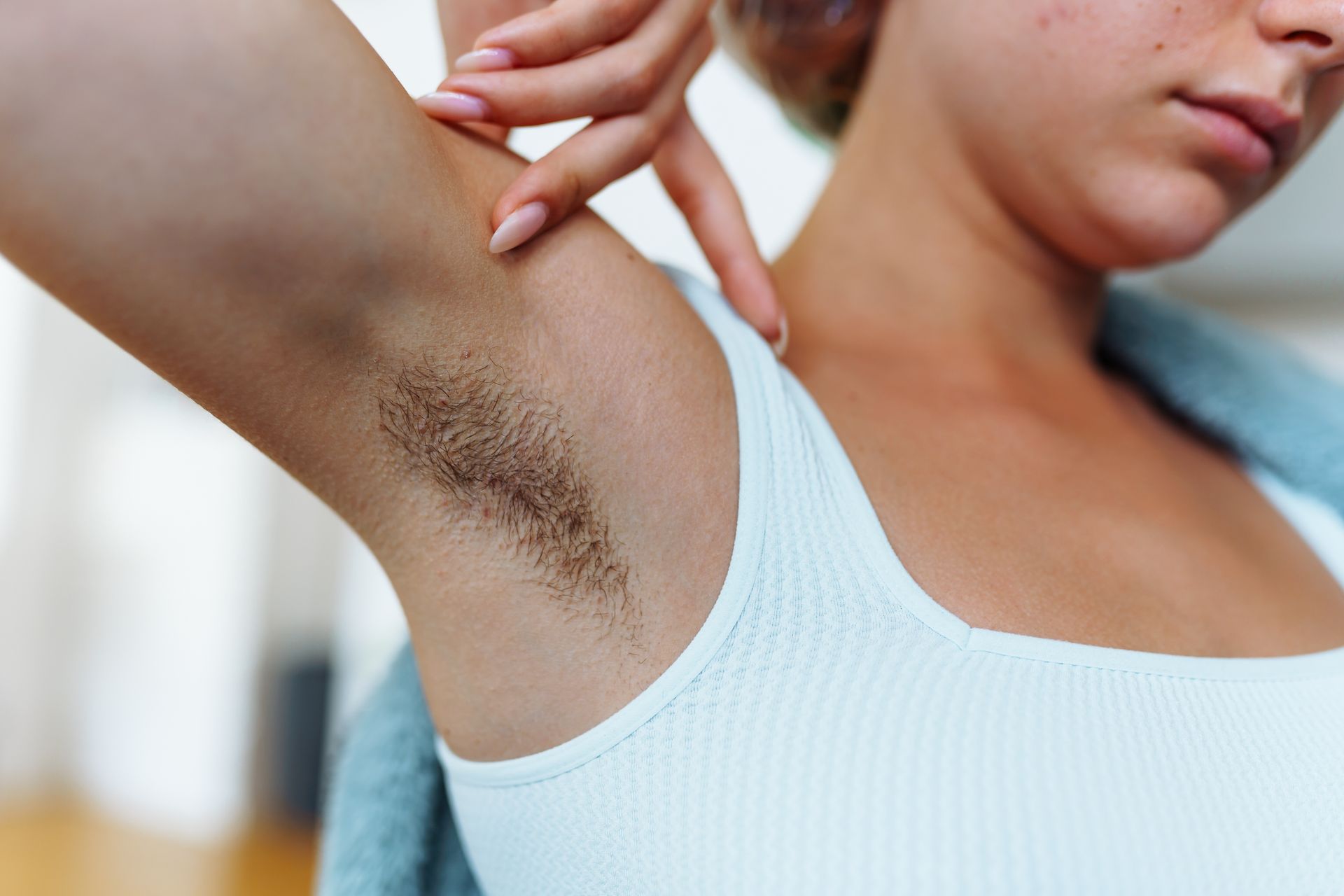 Woman with dark underarm hair, pointing at her armpit. She wears a white tank top.