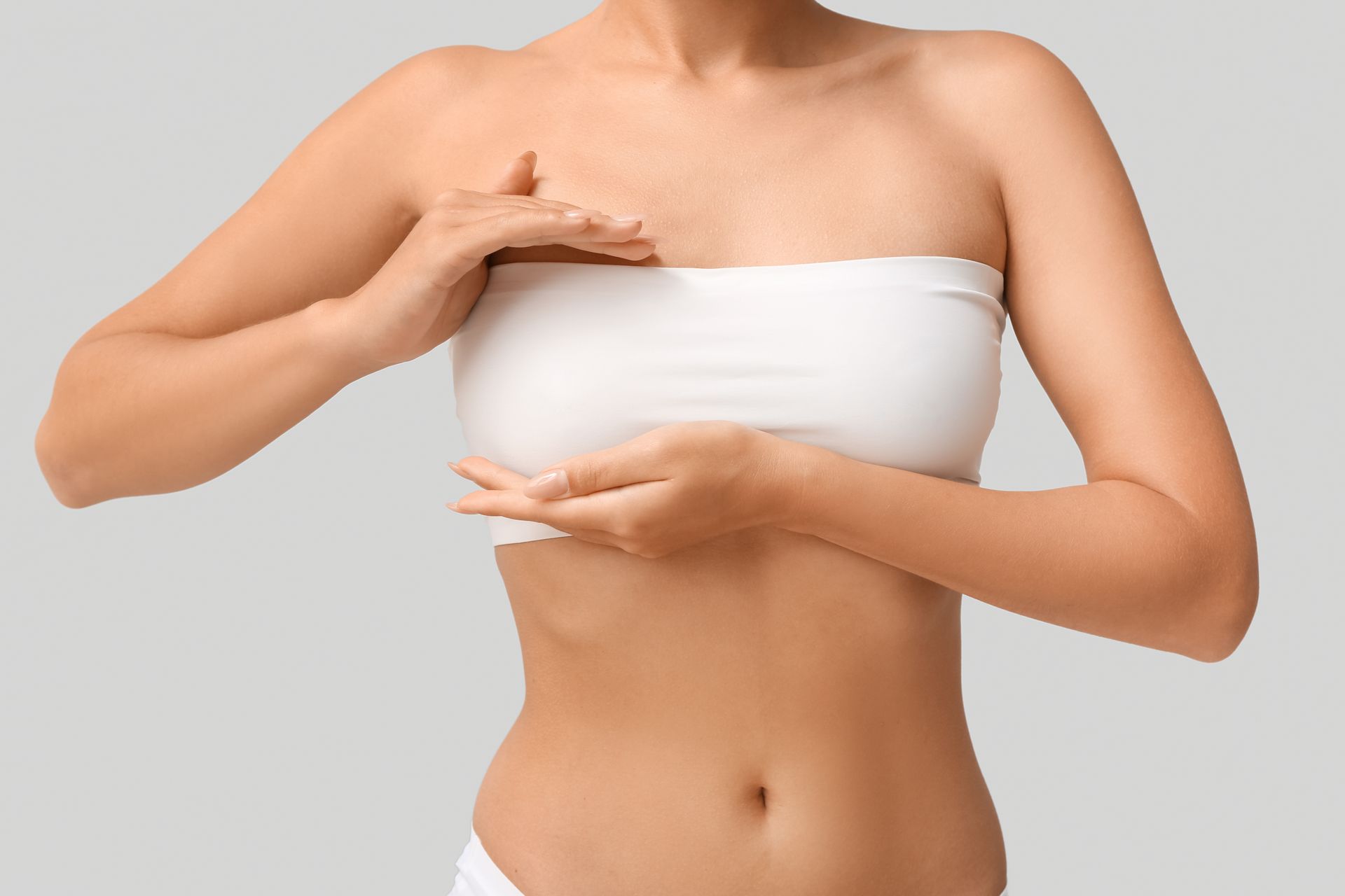 Woman in white bandeau top, holding her breasts. Grey background.