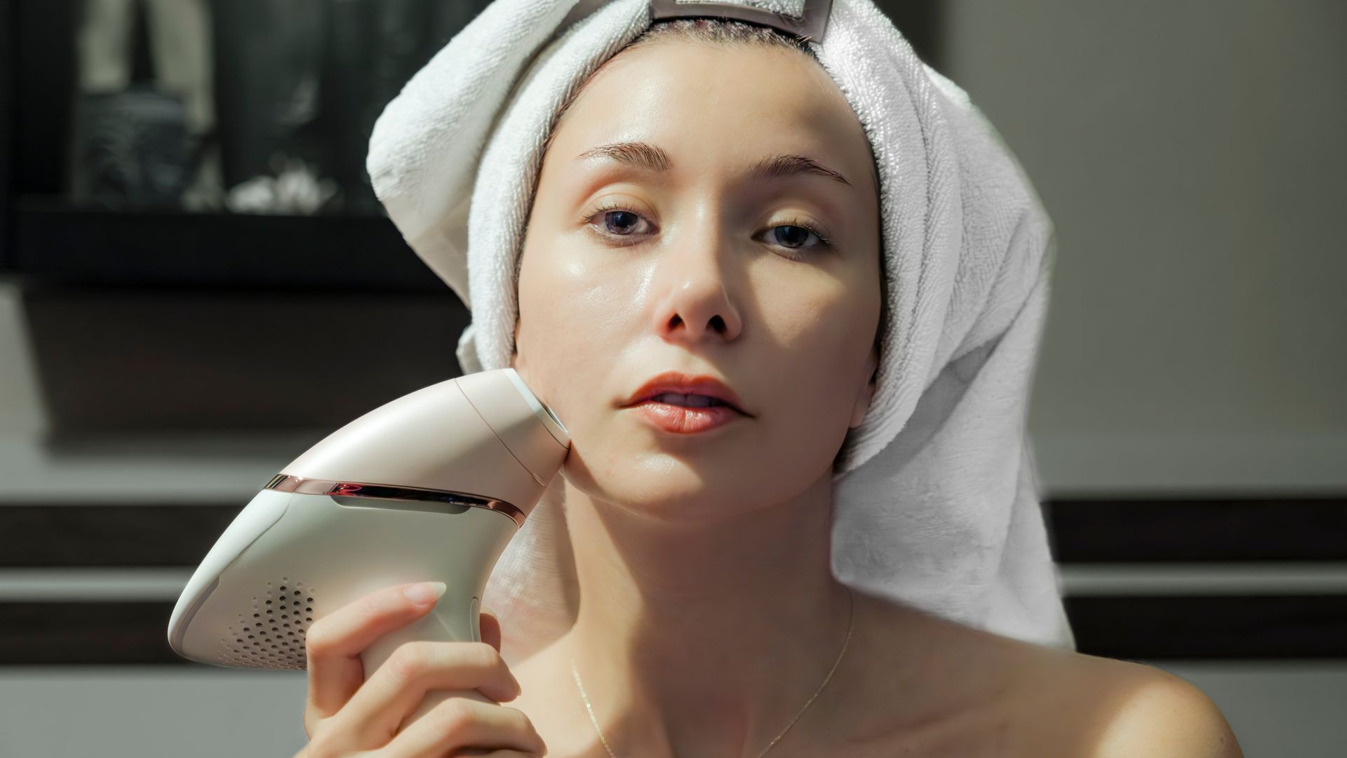 Woman using a handheld device on her neck, wrapped in a white towel; indoor setting.