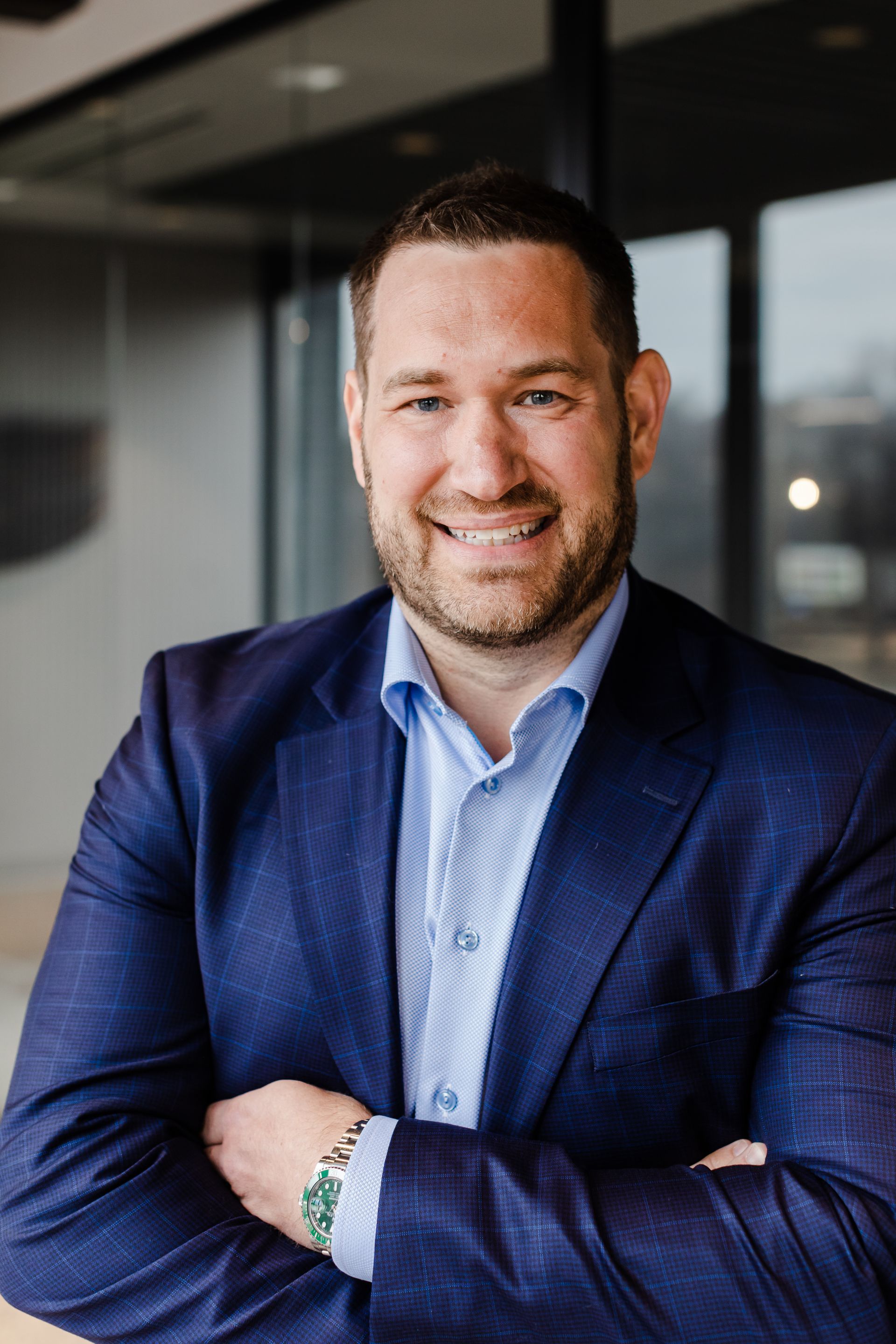 Man in a blue suit smiles, arms crossed, in an office setting.