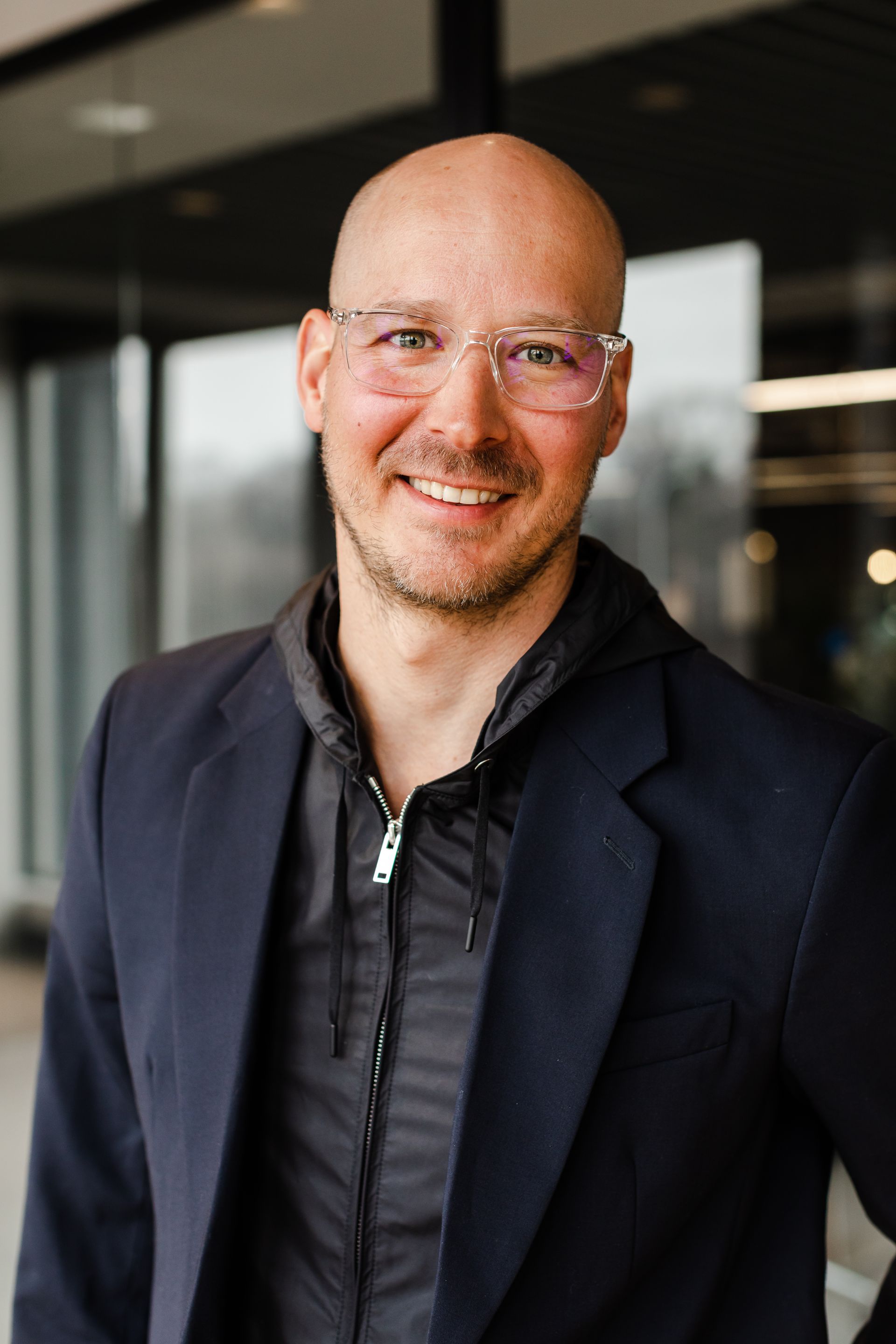 Bald man with glasses, smiling, wearing a black jacket and zipped hoodie, indoors.