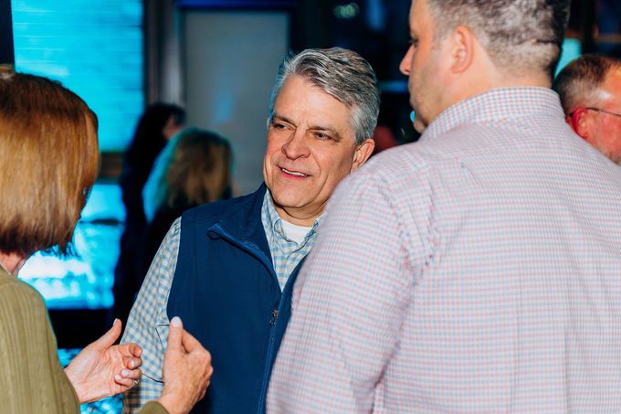 Two men at an event, one eating from a plate, smiling. Others are blurred in the background.
