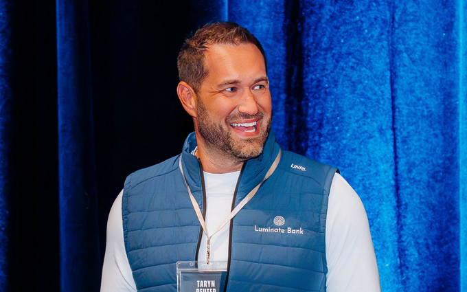 People at a round table, talking. Balloons and food are on the table.  A plaid wall is in the background.