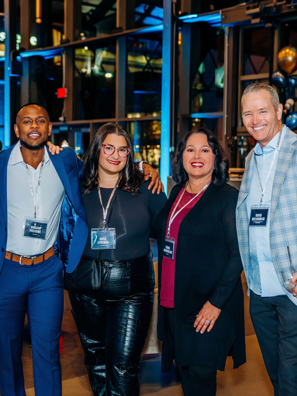 Man in cap and glasses smiles, arms crossed, talking to a woman. Balloons and neon sign in the background.