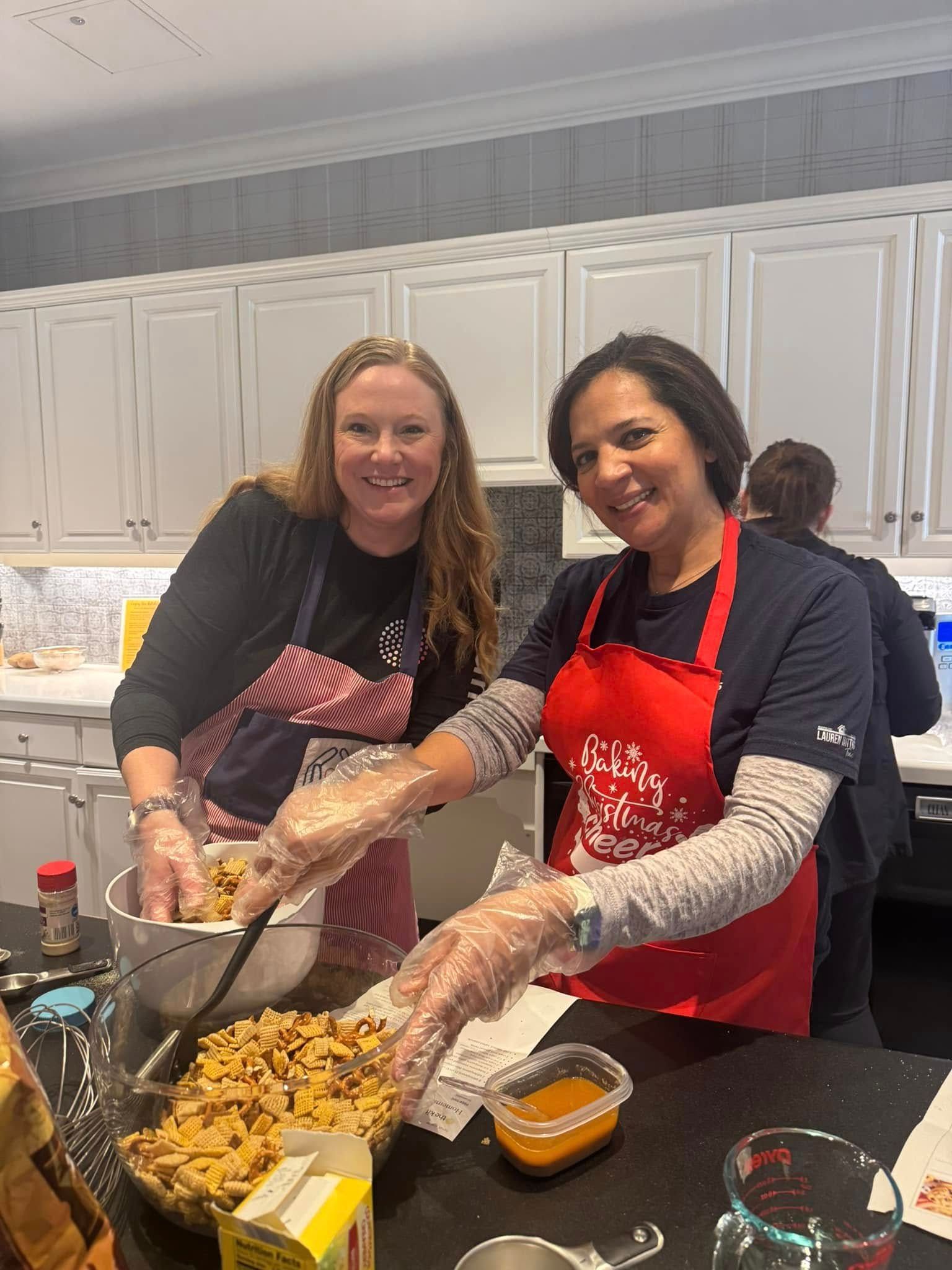 Two women in a kitchen, wearing aprons and gloves, mixing food in a bowl, smiling.