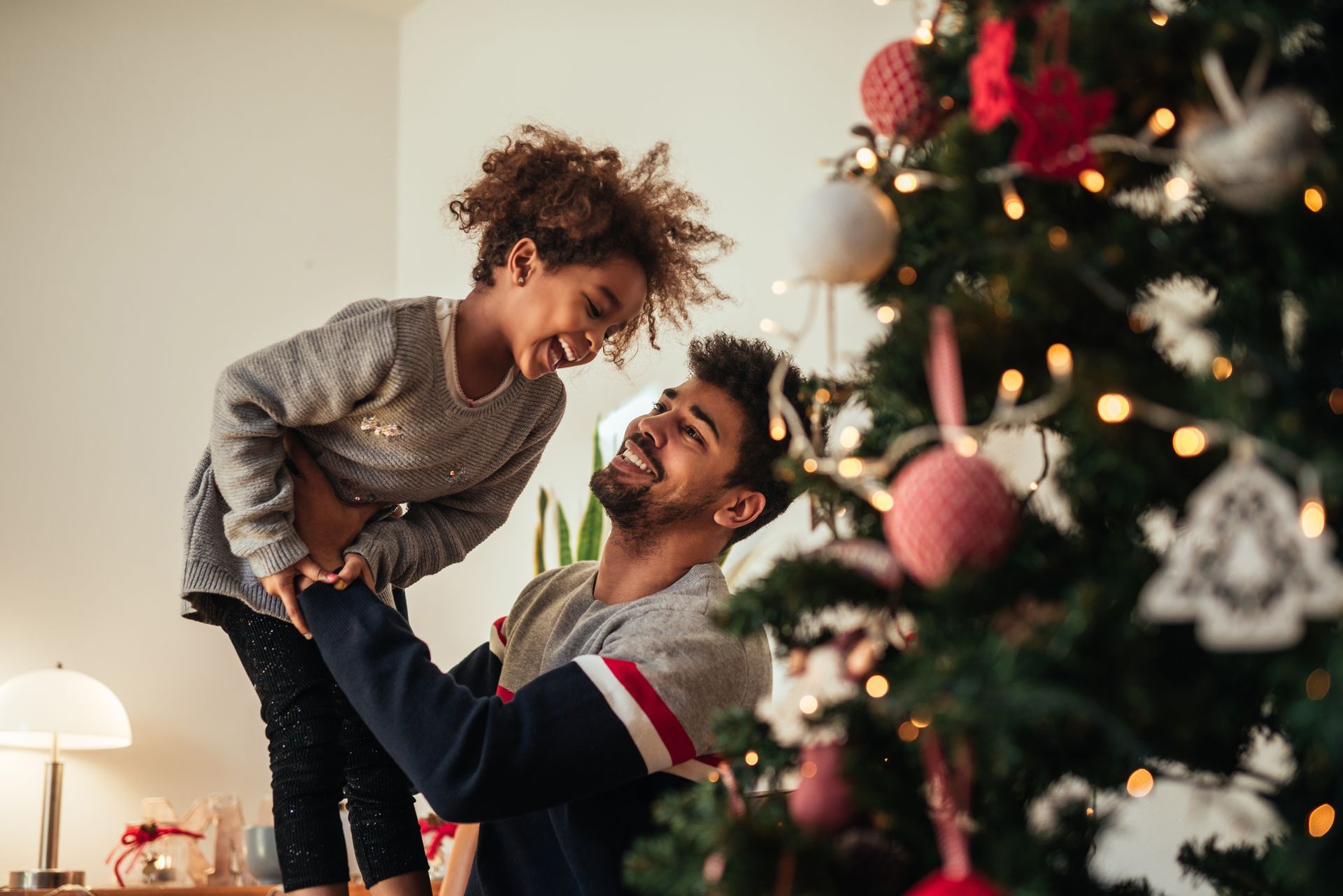 Man and child decorating a Christmas tree, smiling and laughing. Lit tree, ornaments, indoors.