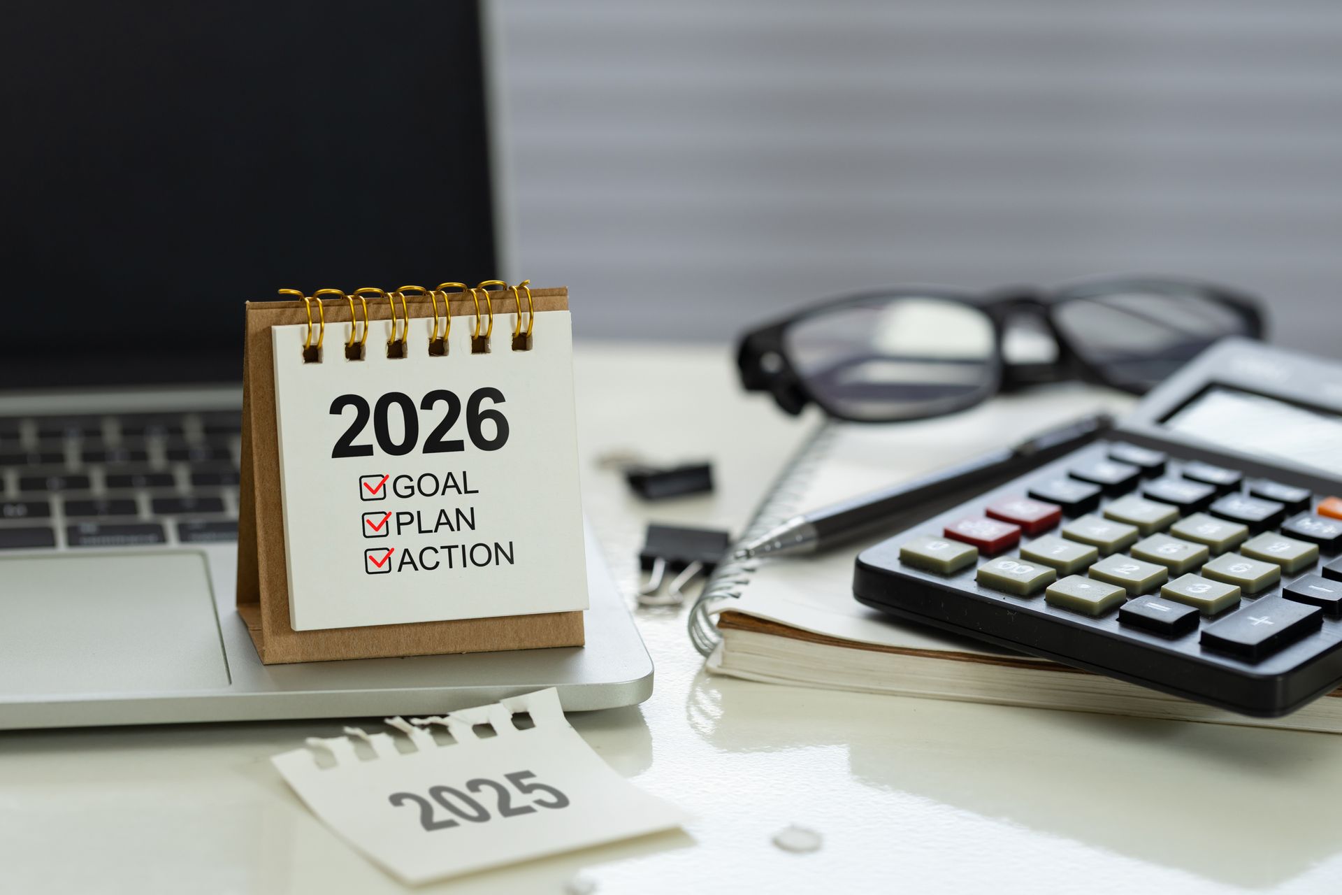 Calendar with 2026, checklist for goal, plan, action; laptop, calculator, glasses, and notepad on a desk.