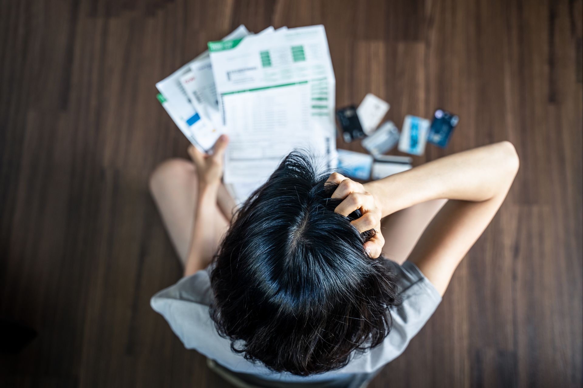 Person sitting, stressed, holding bills and credit cards, on wooden floor.