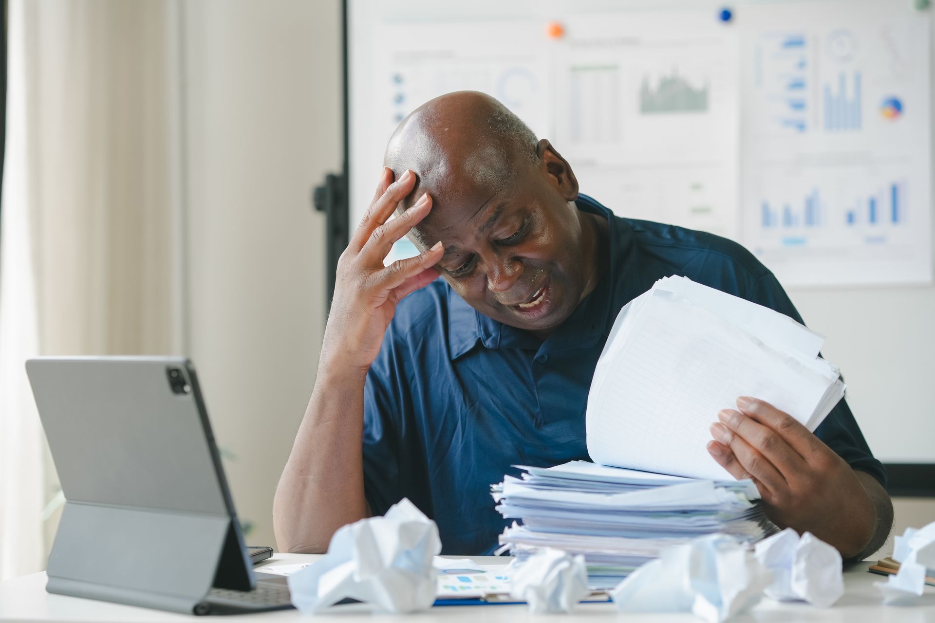 Man stressed at desk, holding papers, with tablet and crumpled paper nearby.