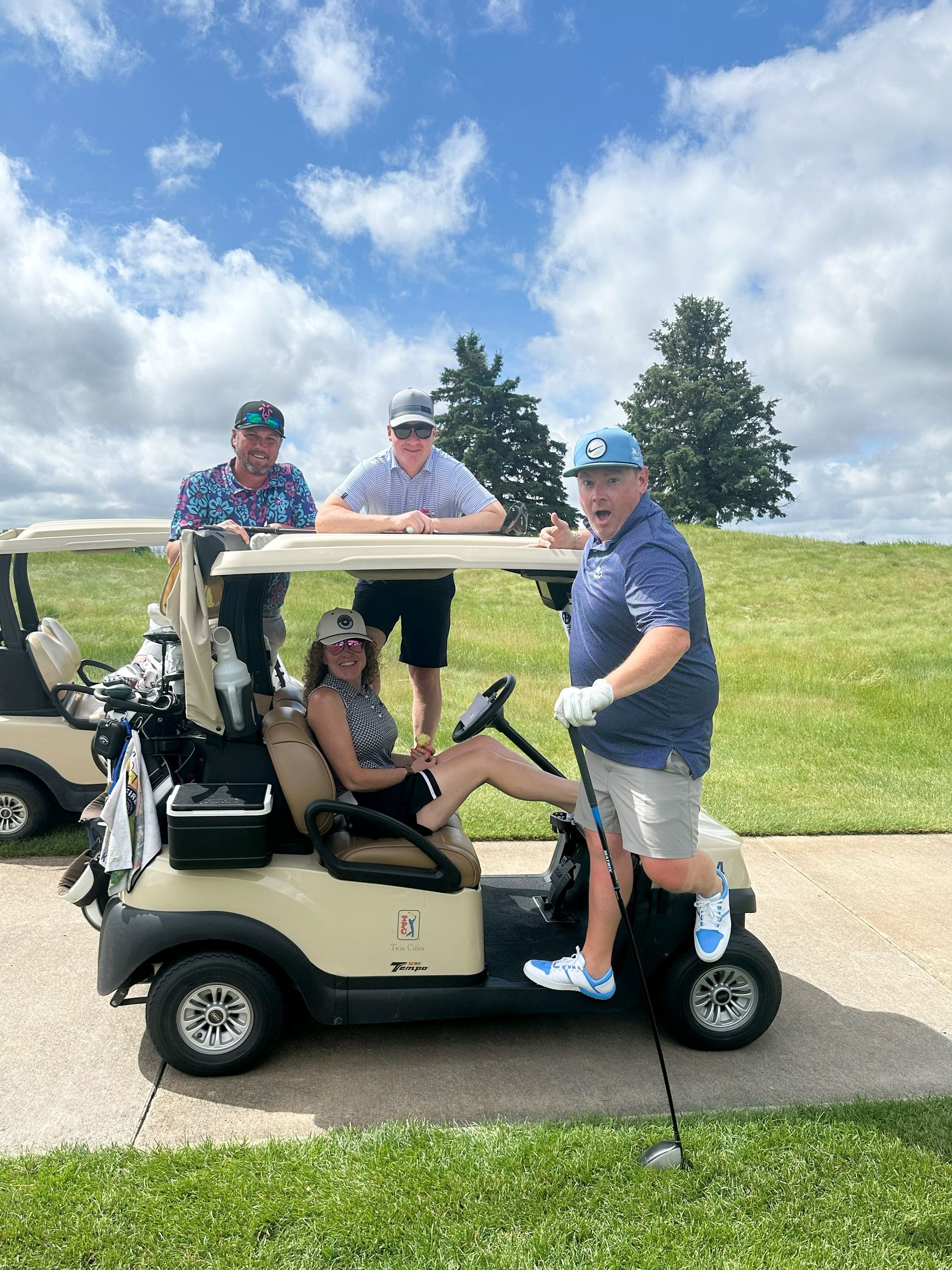 Four people smile on a golf cart, one driving. Sunny day, blue sky.