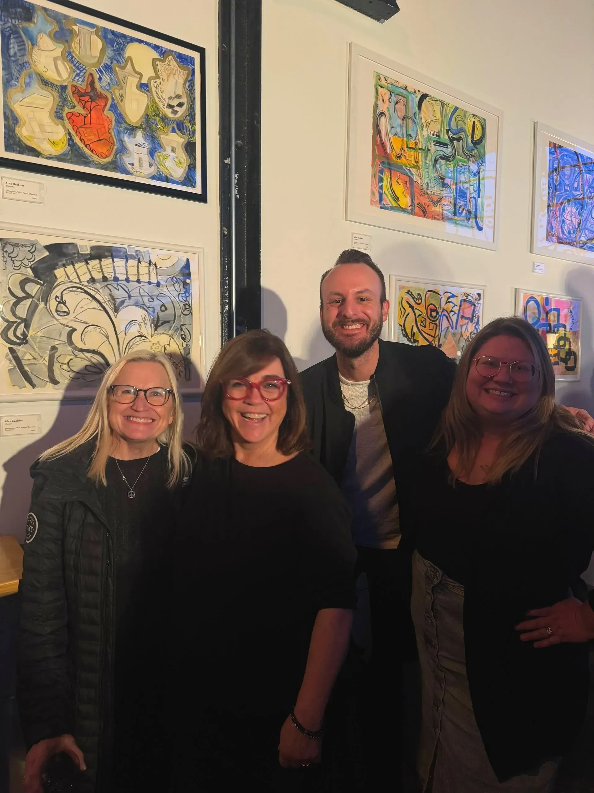 Four people pose in front of framed art. People smile at the camera, against a white wall.