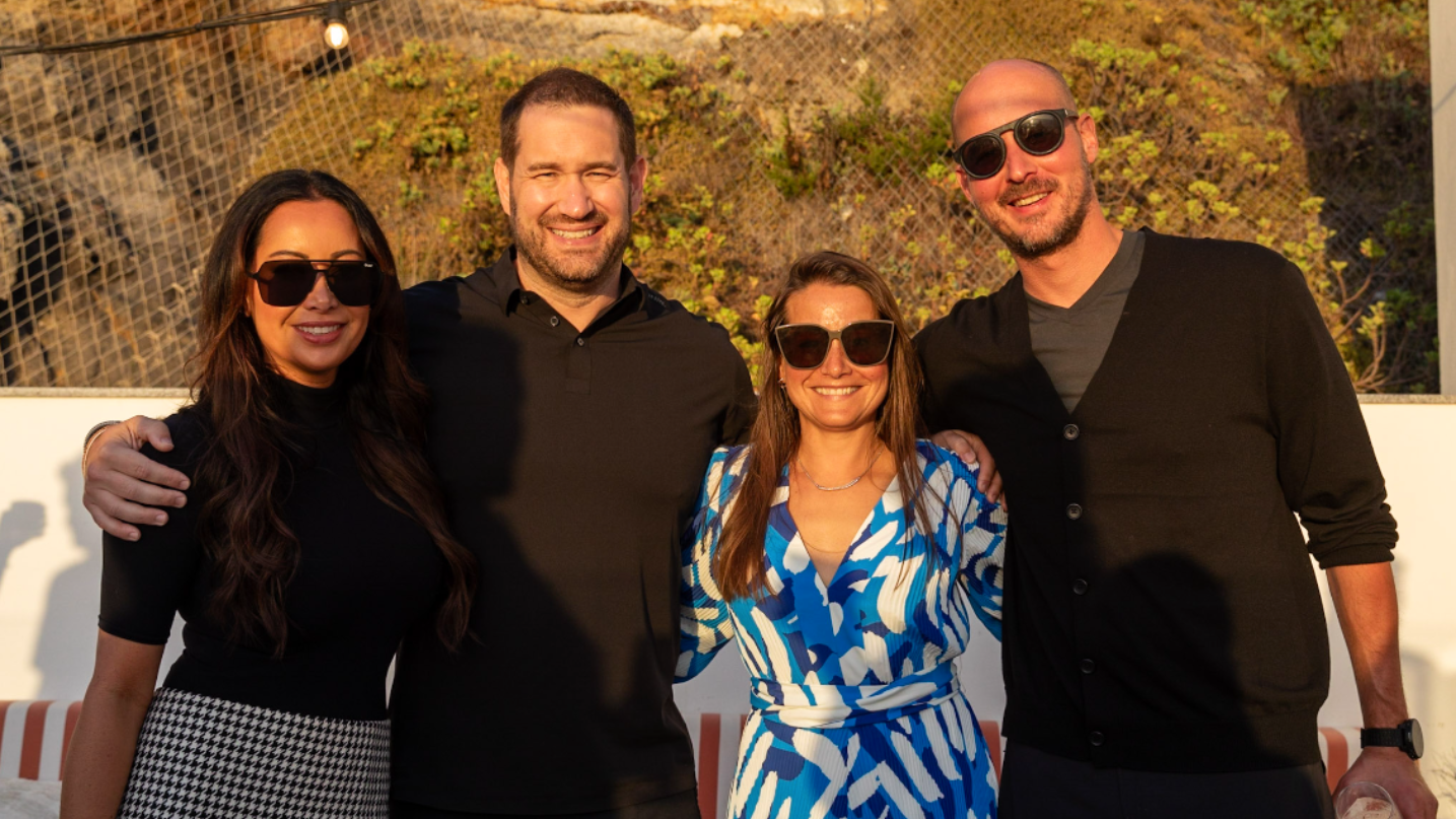 Group of people in life vests on a beach, smiling, with ocean and a banana boat in the background.