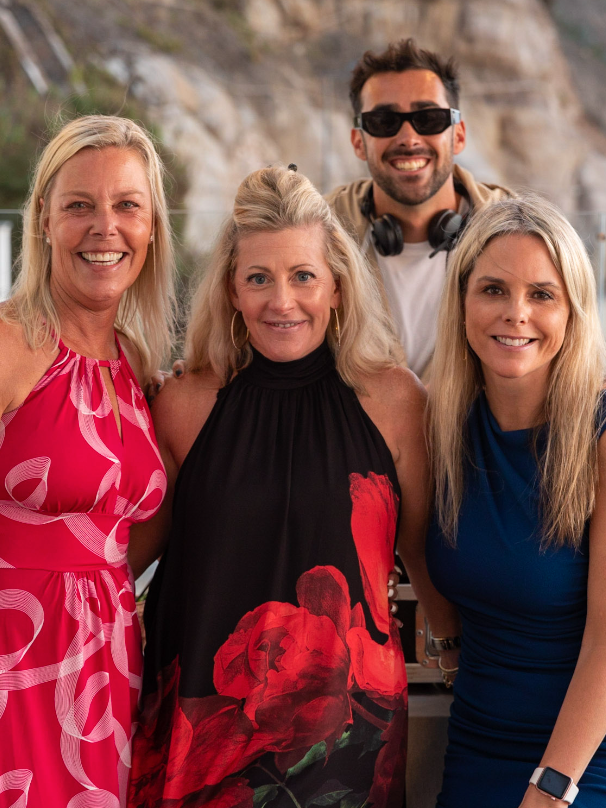 Two women smiling and embracing at a bar; one wears pink, the other a blue blazer.