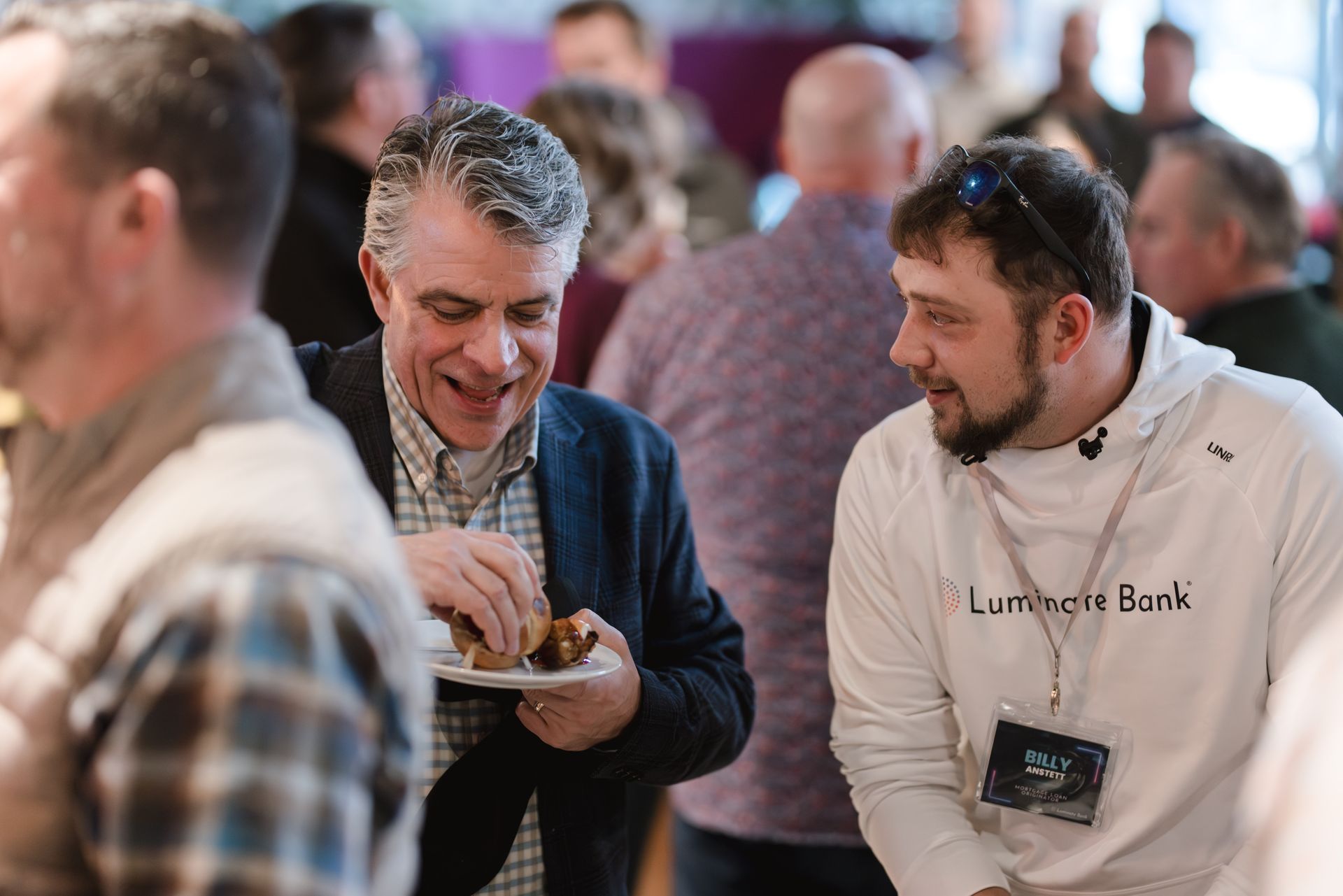 Two men at an event, one eating from a plate, smiling. Others are blurred in the background.