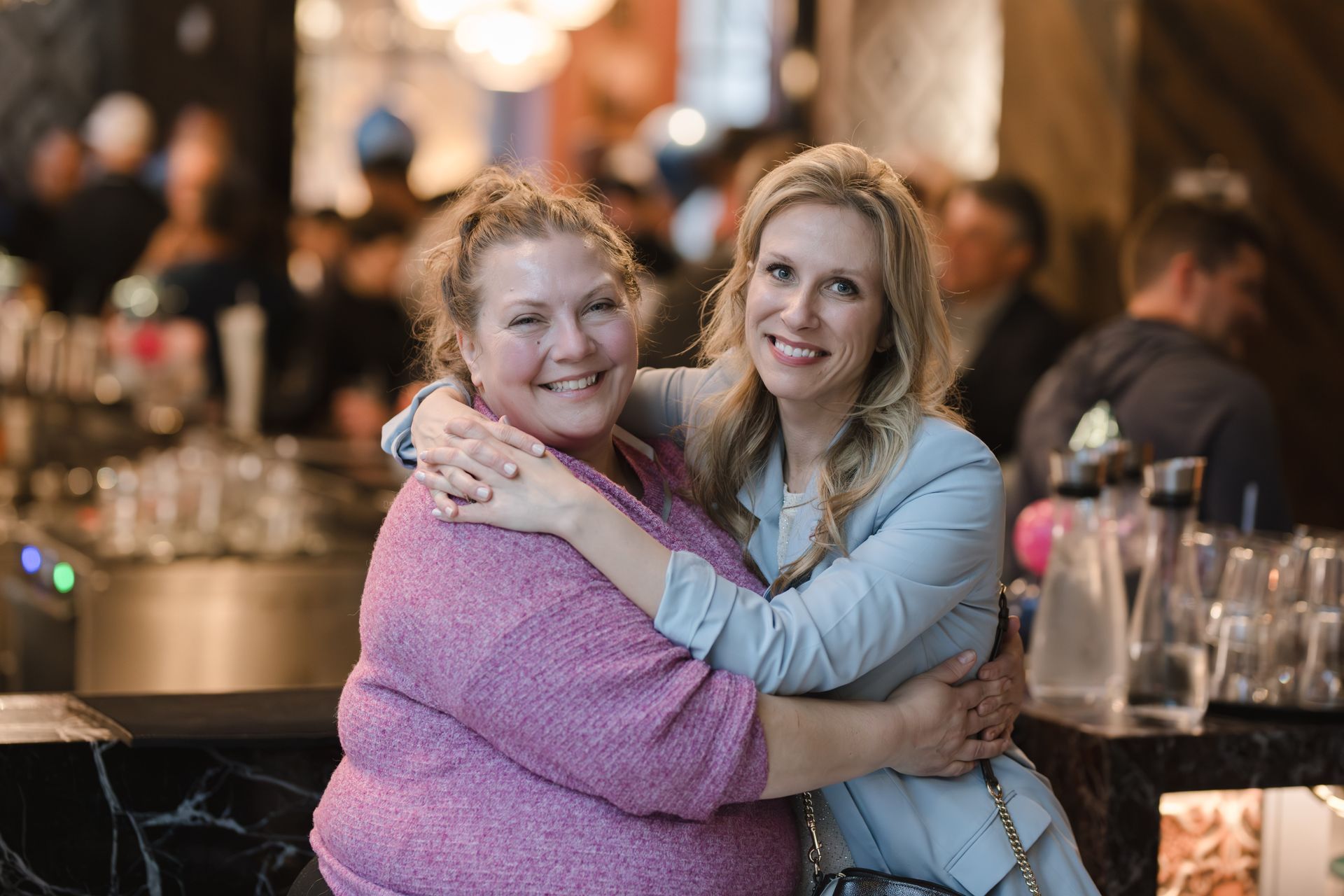 Two women smiling and embracing at a bar; one wears pink, the other a blue blazer.