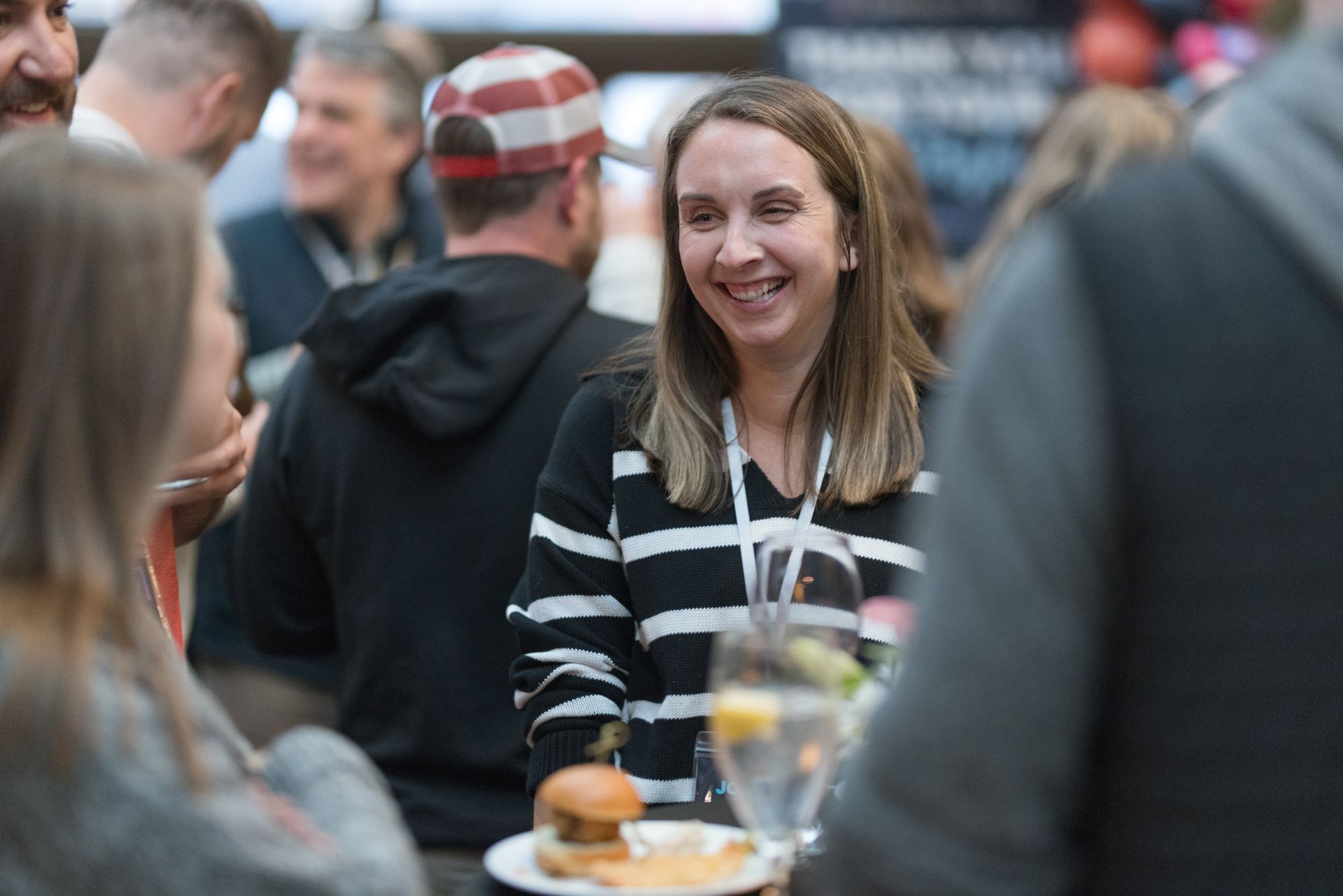 Woman smiles at event, wearing striped sweater, near food and drinks. Surrounded by people in a social setting.