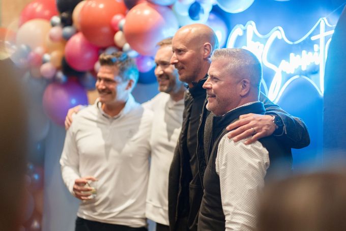 Woman smiles at event, wearing striped sweater, near food and drinks. Surrounded by people in a social setting.