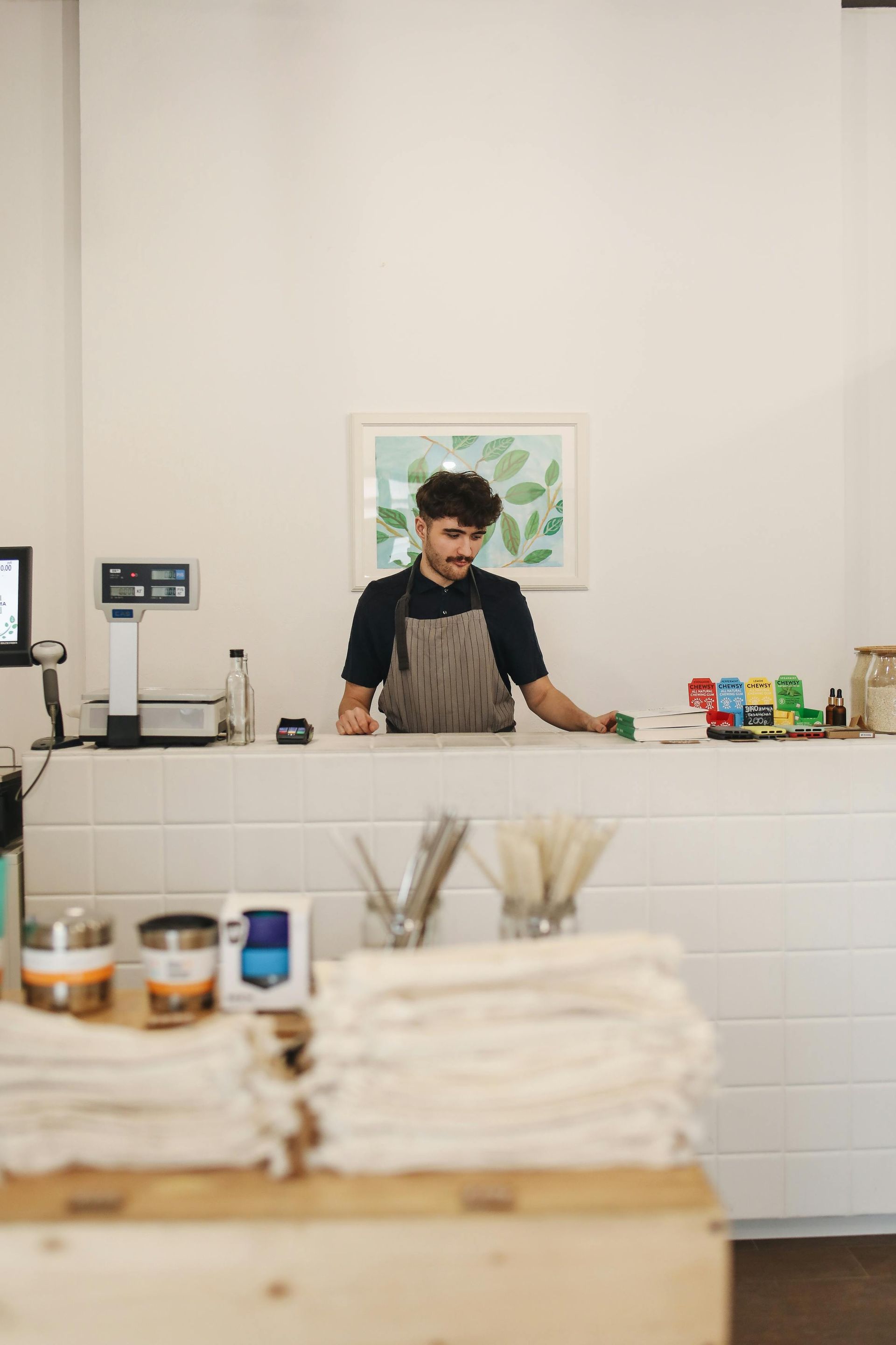 Man in apron stands at a store counter, looking down. Behind him is a small painting.