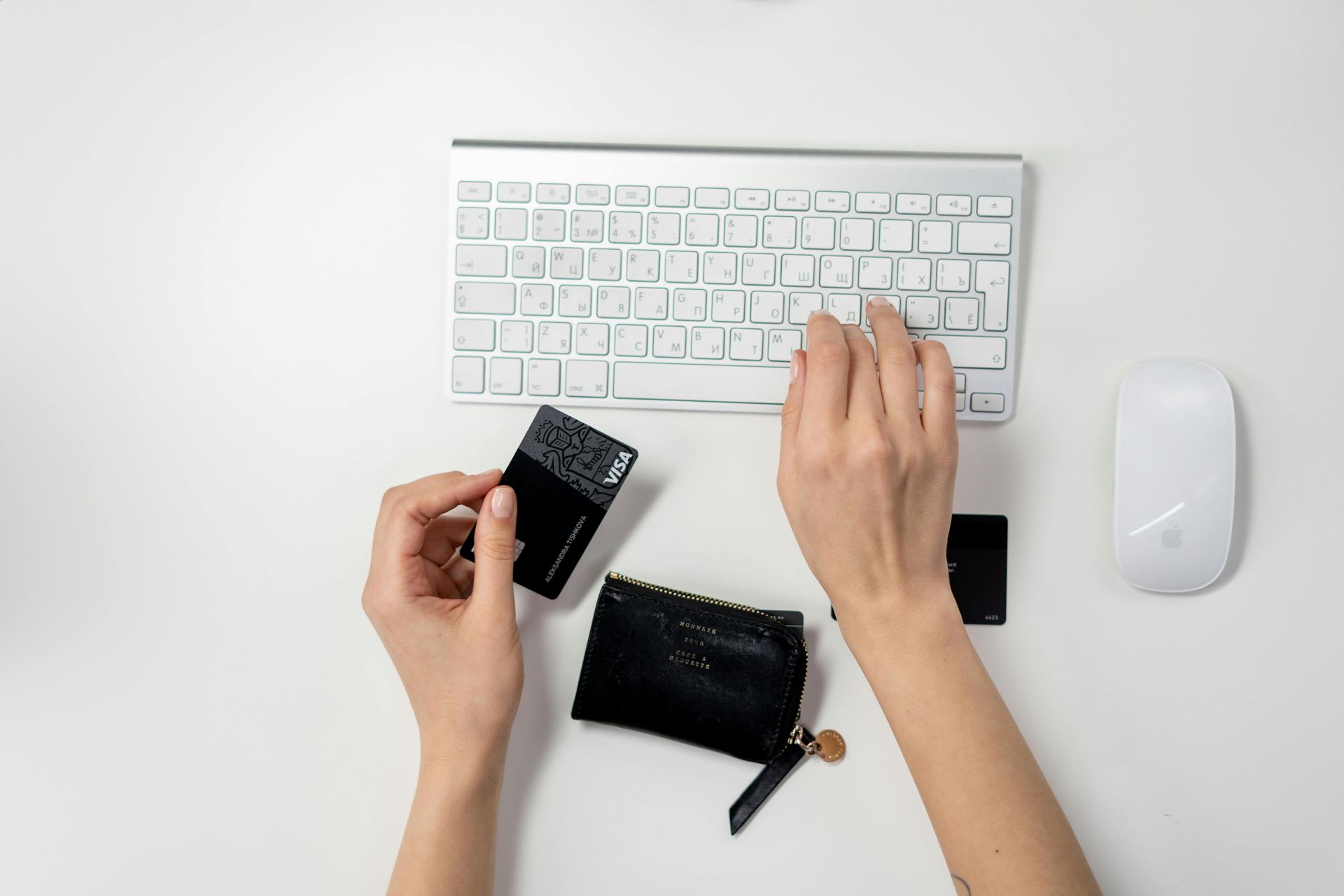 Person using a keyboard and mouse, holding a credit card next to a wallet on a white surface.