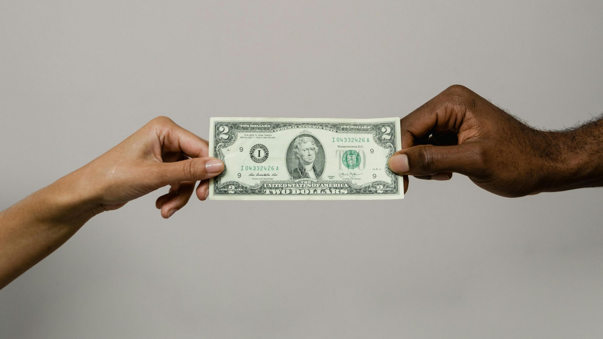 Two hands, one light-skinned and one dark-skinned, holding a one-dollar bill against a gray backdrop.