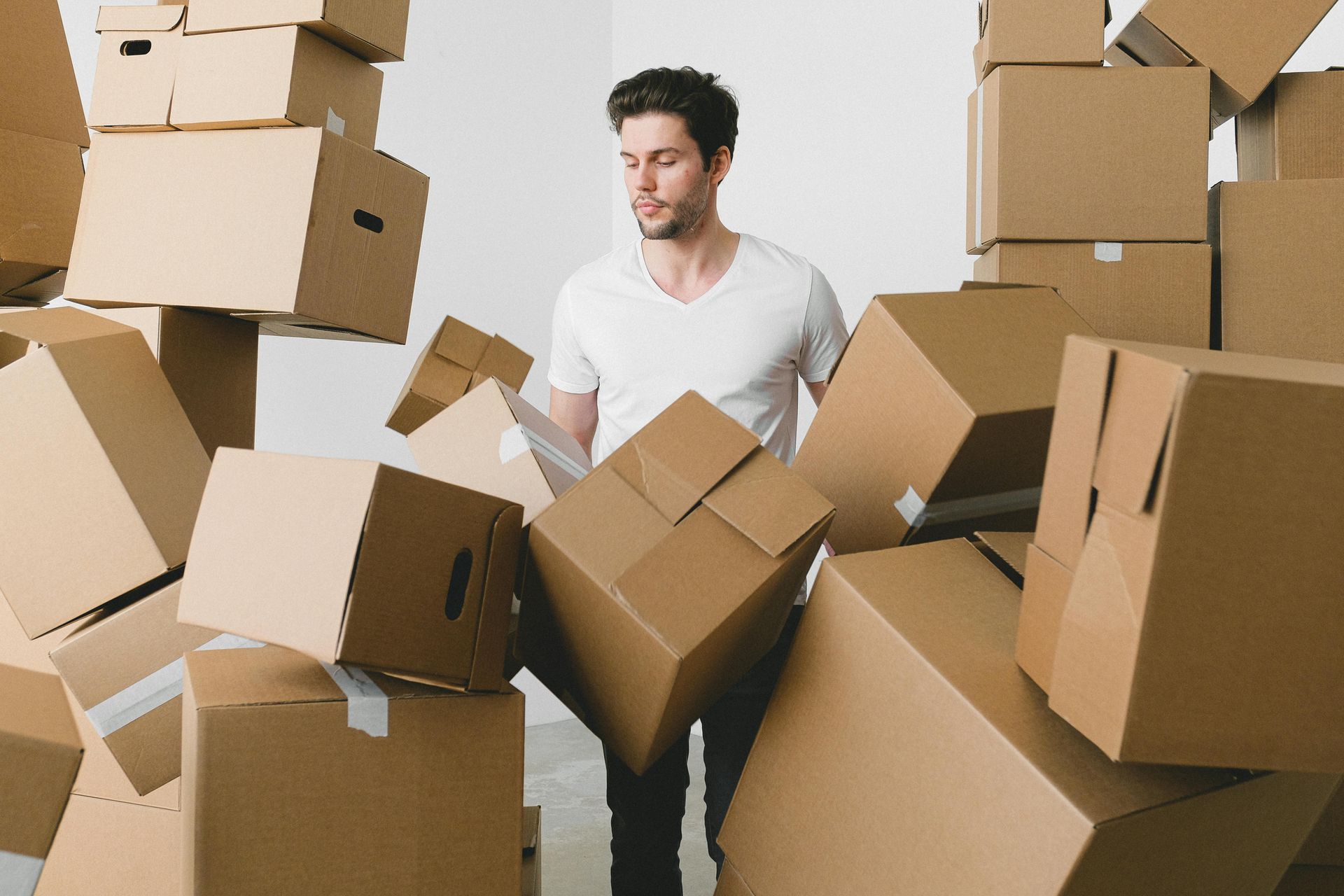 Man surrounded by cardboard boxes, holding one, with a concerned expression in a room.