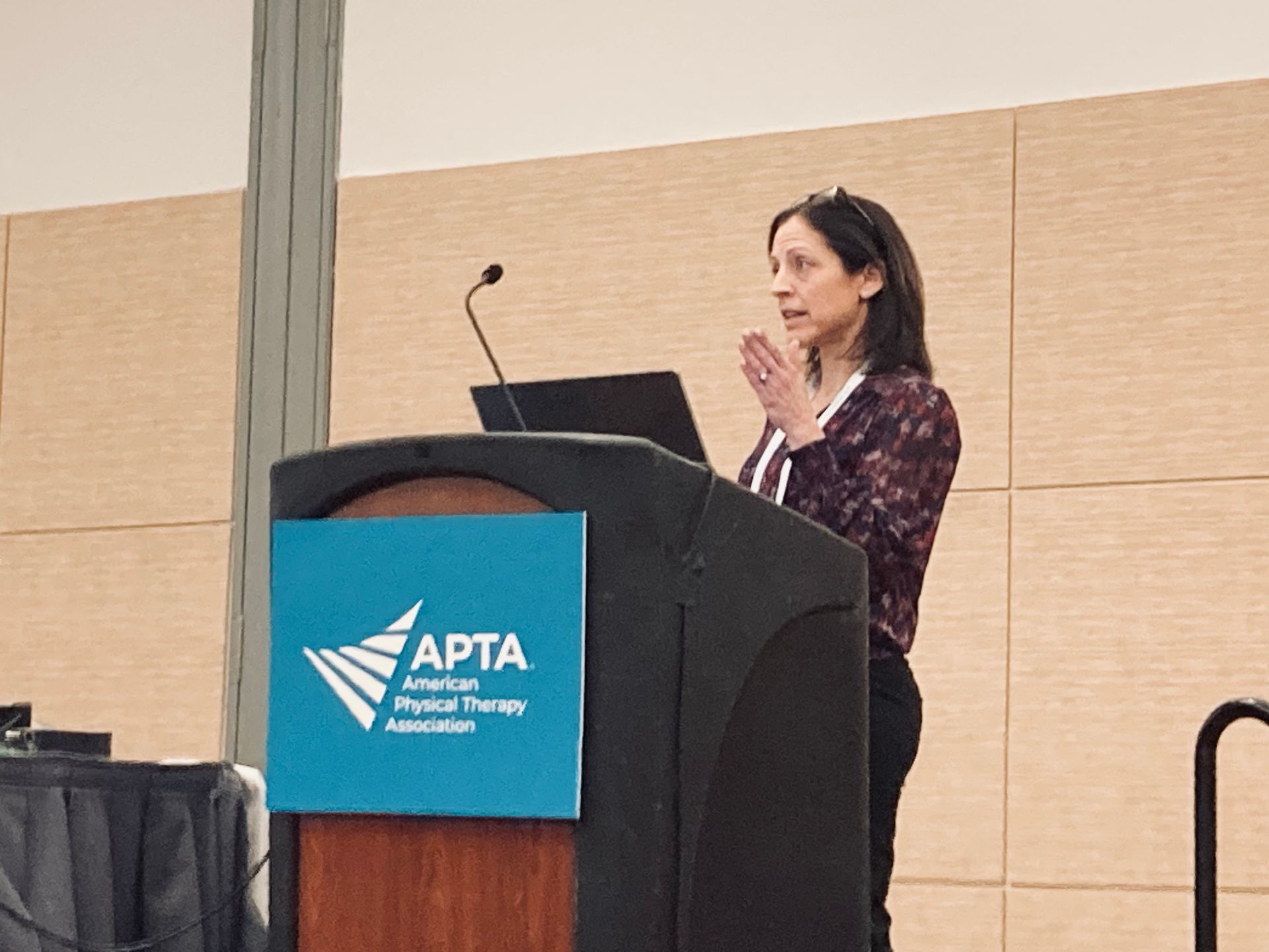 Woman presenting in a classroom. She's standing behind a podium with a screen showing a presentation.