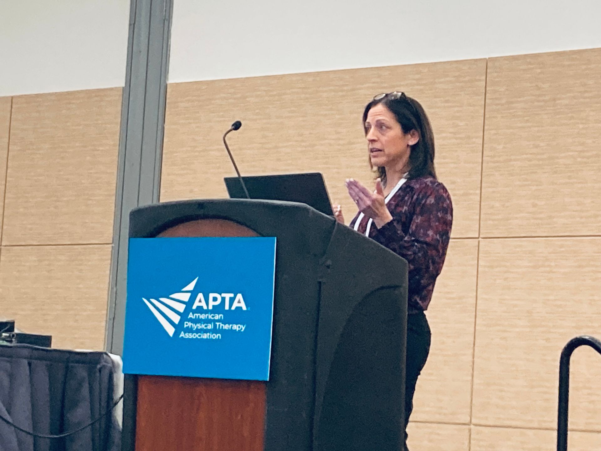 Woman speaking at a podium with APTA logo. She is gesturing, wearing a patterned top and dark pants. Conference setting.
