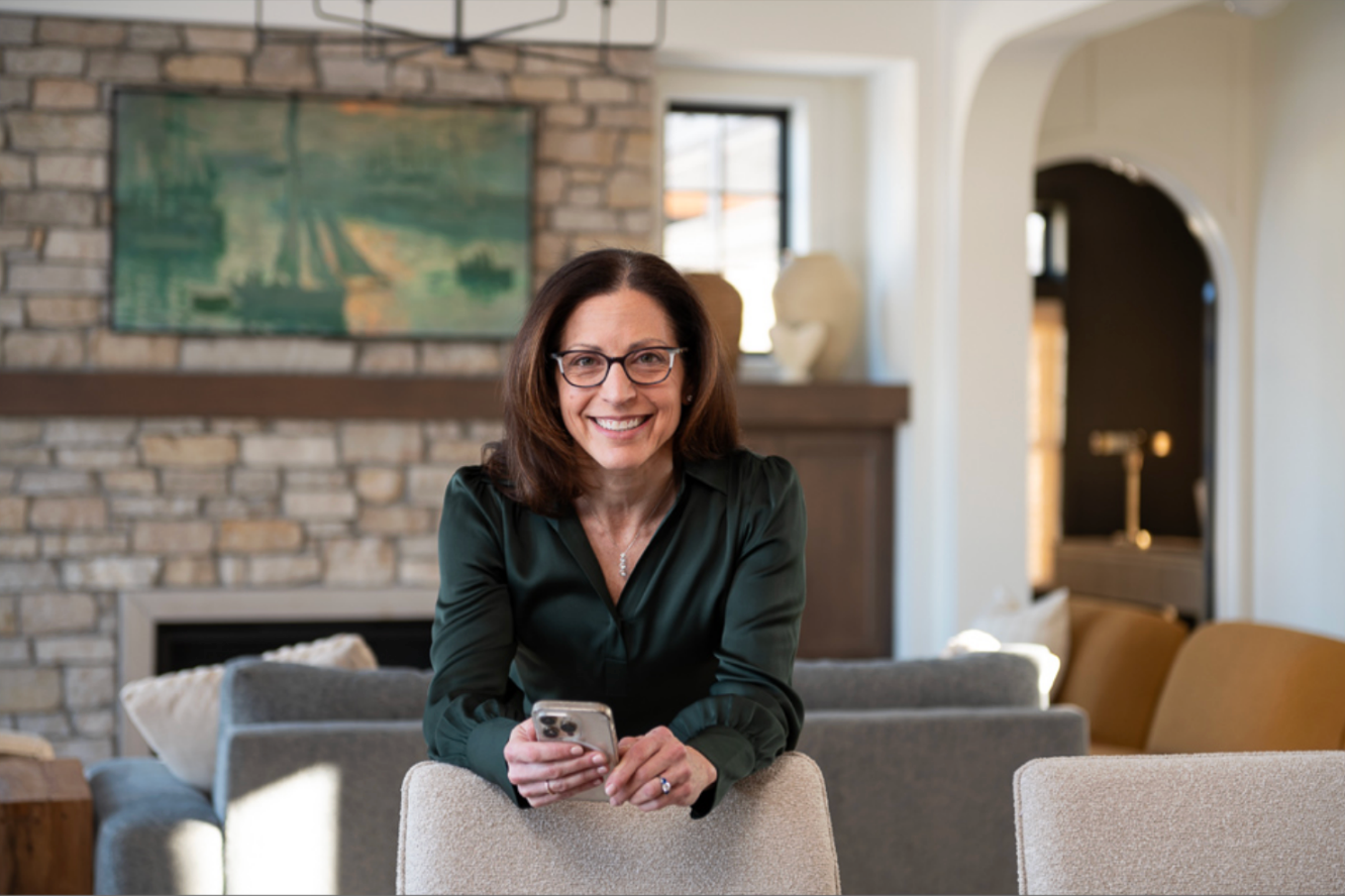 Woman in glasses, dark green top, leaning on chair, smiling in front of fireplace and sofa.