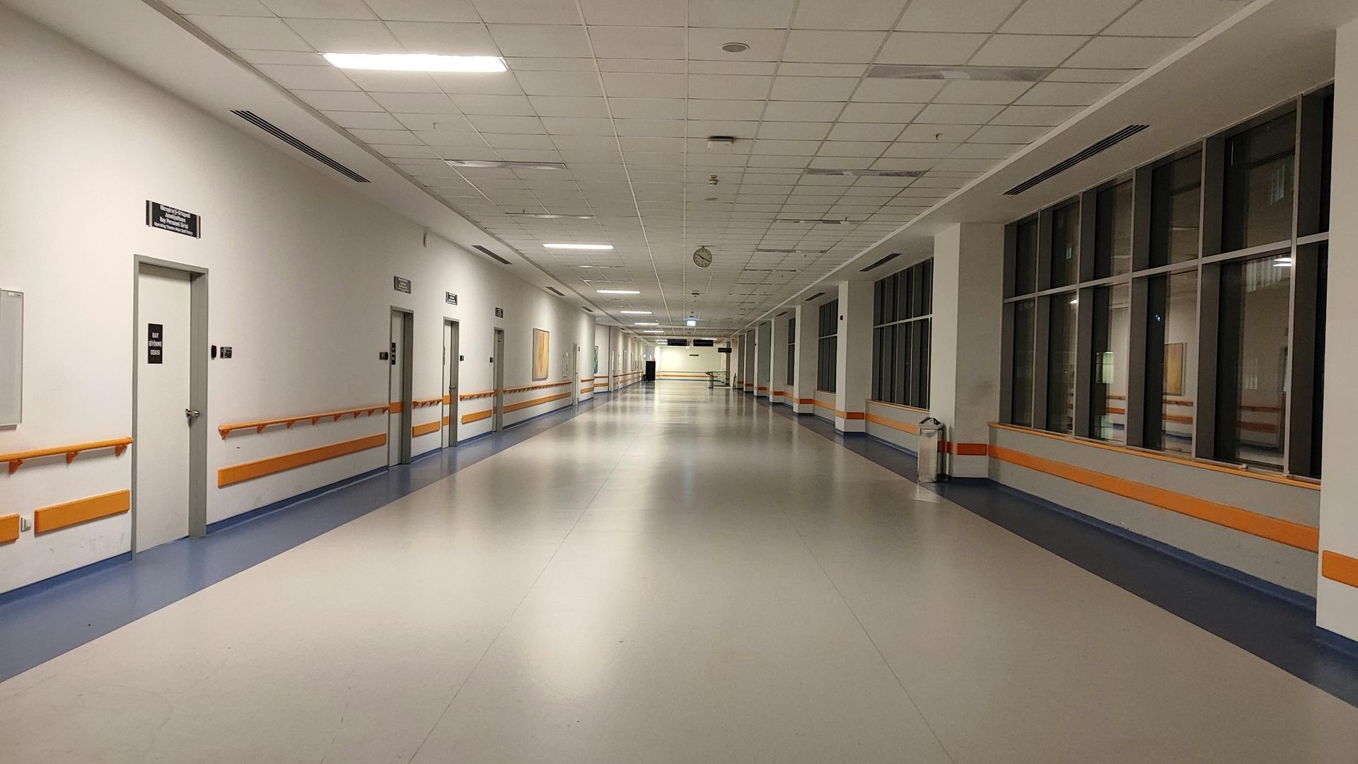 Empty hospital hallway, white walls, blue and grey floor, doors on the left, windows on the right.
