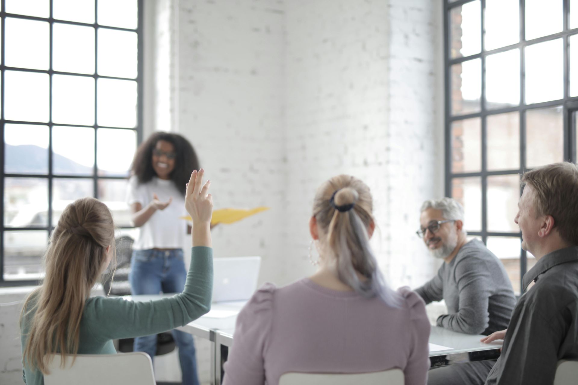 People in a meeting, one woman raising her hand, another speaking. Bright, sunny room.