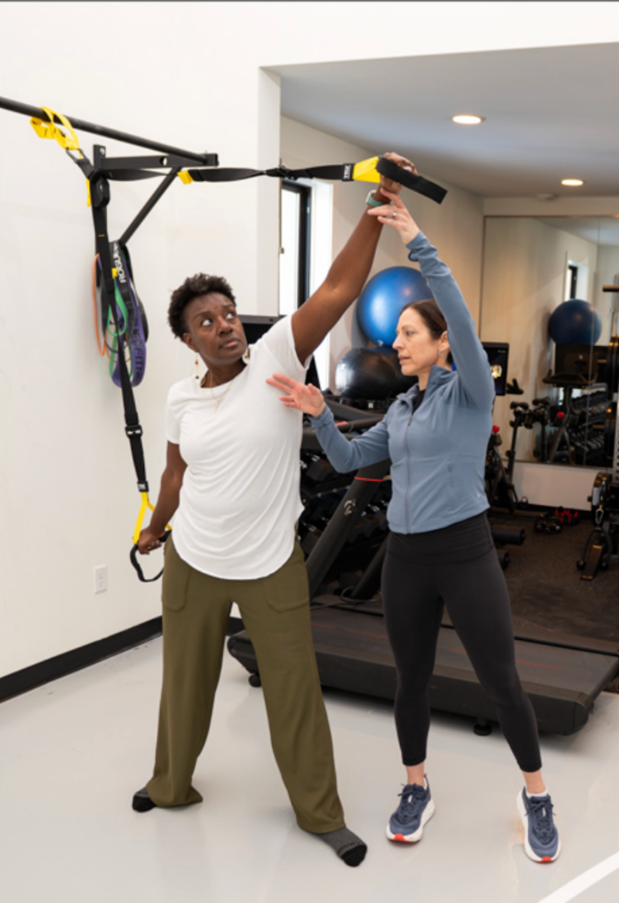 Woman in white shirt using TRX with instructor in a gym setting. Instructor is guiding.