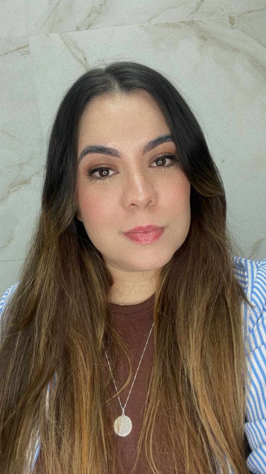 Mulher de longos cabelos castanhos olha para a câmera. Ela está em uma sala, vestindo uma blusa marrom, uma camisa azul e branca e um colar.