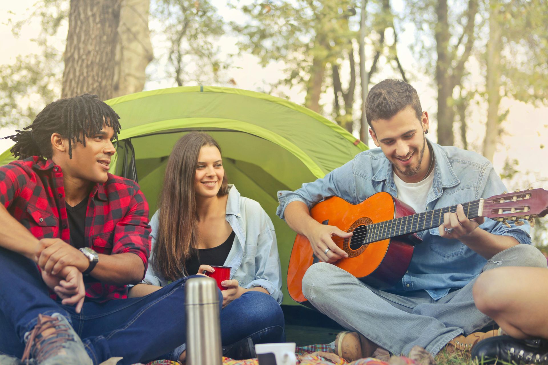 Três amigos acampando, um deles tocando violão. Barraca ao fundo, árvores e luz do sol.