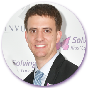 A man in a suit and tie is smiling in front of a sign that says solving cancer