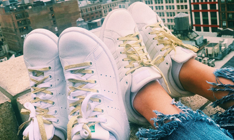 A person wearing a pair of white sneakers with gold laces