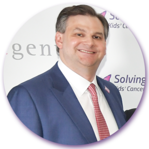 A man in a suit and tie is smiling in front of a sign that says solving kids cancer