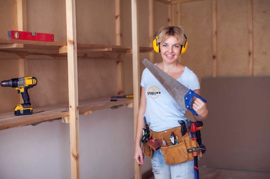 A drywall company female contractor wearing a white tank top and tool belt holds a saw while standing in a room with wooden wall framing. She is working on a residential project near S 27th Ave, and W Shaughnessey Rd, Ahwatukee Foothills, AZ.