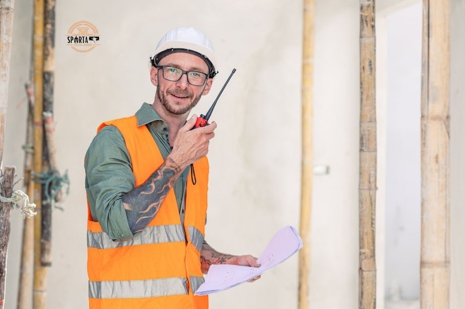 A smiling drywall contractor wearing protective equipment holds a walkie-talkie while standing among wooden wall studs and framing in a drywall project. He is working near N Harbour Town CT, and W Wolf Run Dr, Anthem, AZ.