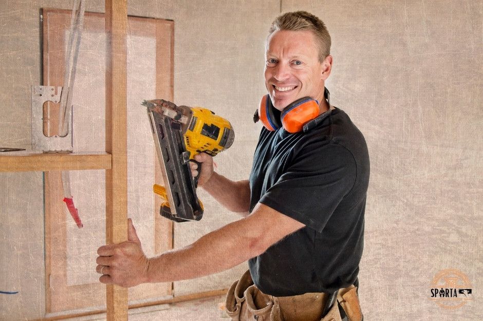 A drywall company worker in a black shirt and tool belt uses a screw gun to install drywall panels on wooden wall framing in a residential construction site. The man is actively working on the interior walls near W Rio Vista Ln, and S 115th Dr, Avondale, AZ.