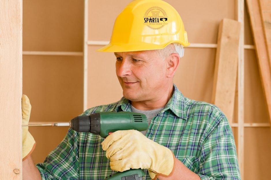 A drywall company worker wearing yellow hard hat and green plaid shirt holds power drill while smiling. Residential construction worker stands in front of wooden wall framing and drywall panels at job site near W Ashley Dr, and S 258th Ave, Buckeye, AZ.