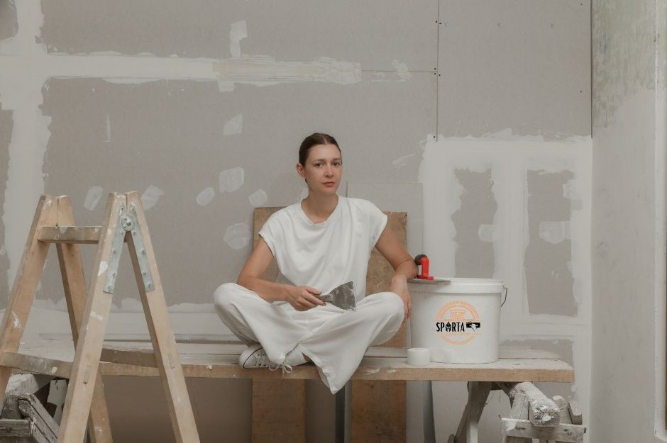 A drywall company worker sits on wooden sawhorse in white clothing, holding a putty knife in a residential construction site, surrounded by unfinished drywall walls with patches. Wooden ladder and supplies visible on floor near E Brooklyn Dr, and S Wetleaf Way, Casa Grande, AZ.