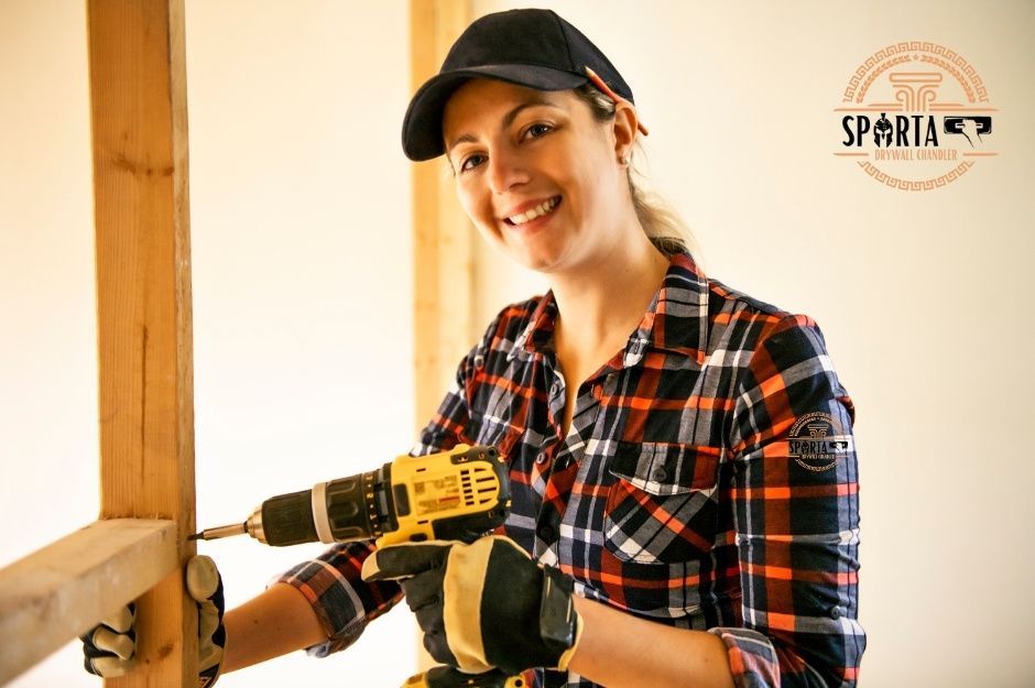 A drywall company worker in a plaid flannel shirt and black cap holds a yellow cordless drill while smiling at the camera. She stands in an unfinished residential construction with white walls and exposed wooden framing visible in the background near W Montebello Ave, and N 184th Dr, Citrus Park, AZ.