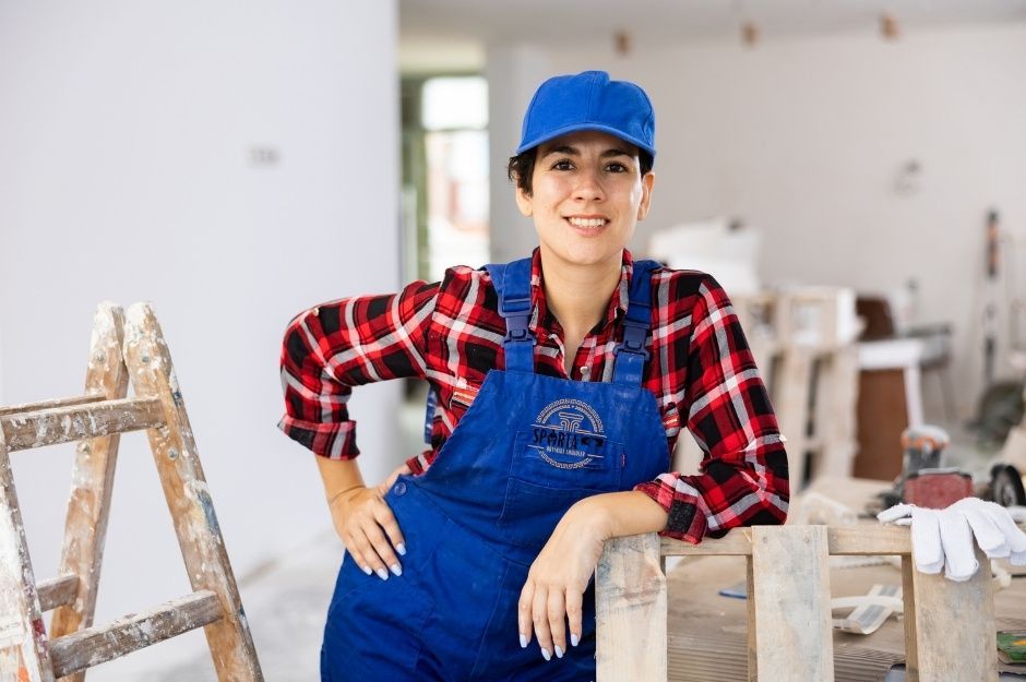 A confident drywall contractor in proper safety gear—blue hat and work overalls over a red plaid shirt—leans casually against a wooden ladder in a drywall project. The drywall project is near S Butte St, and E Third Aly, Dewey–Humboldt, AZ.