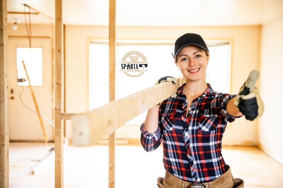 A drywall company professional in a red plaid shirt and a hat holds a piece of wood while giving a thumbs up in an unfinished room with exposed wooden framing and bright lighting. She appears confident and ready for residential construction work near W Charter Oak Rd, and N 123rd Dr, El Mirage, AZ.