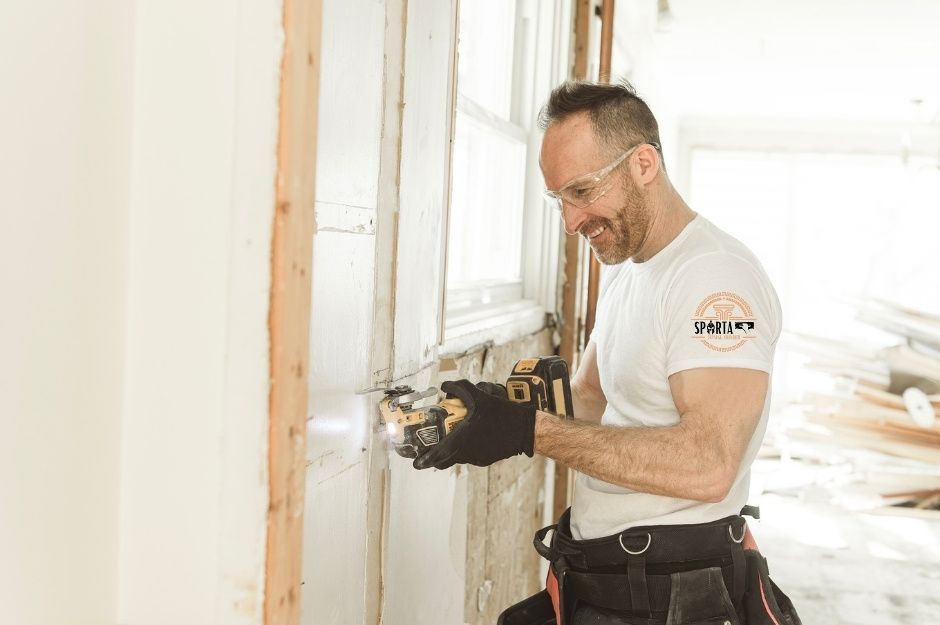 A drywall contractor in a white t-shirt uses a power drill while working on drywall installation near wooden framing and a window. This drywall company professional is completing a project near Firerock, Fountain Hills, AZ.