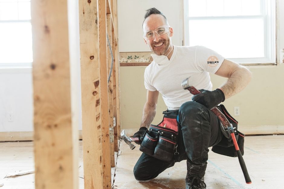 A drywall contractor in a white t-shirt kneels while working with drywall installation tools near wooden framing in a bright construction space. This drywall professional is completing a project near W Orchard Way, and N Summerfield Dr, Gilbert, AZ.