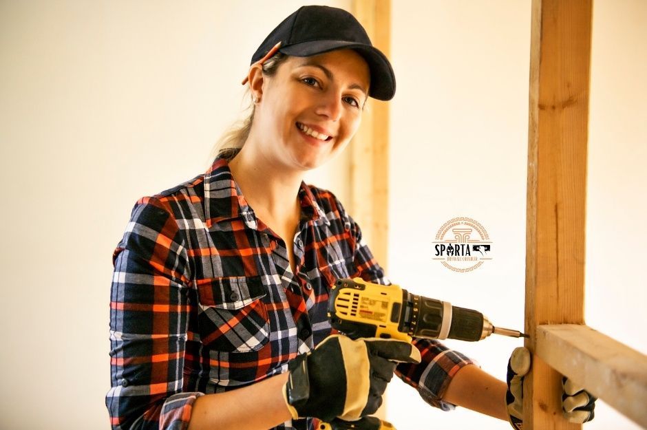 A drywall company worker smiles while holding a yellow power drill in an indoor residential construction setting. She wears a plaid flannel shirt and black baseball cap, standing in what appears to be an unfinished room with exposed framing near E Calle Vahi, and S Calle Tomi, Guadalupe, AZ.
