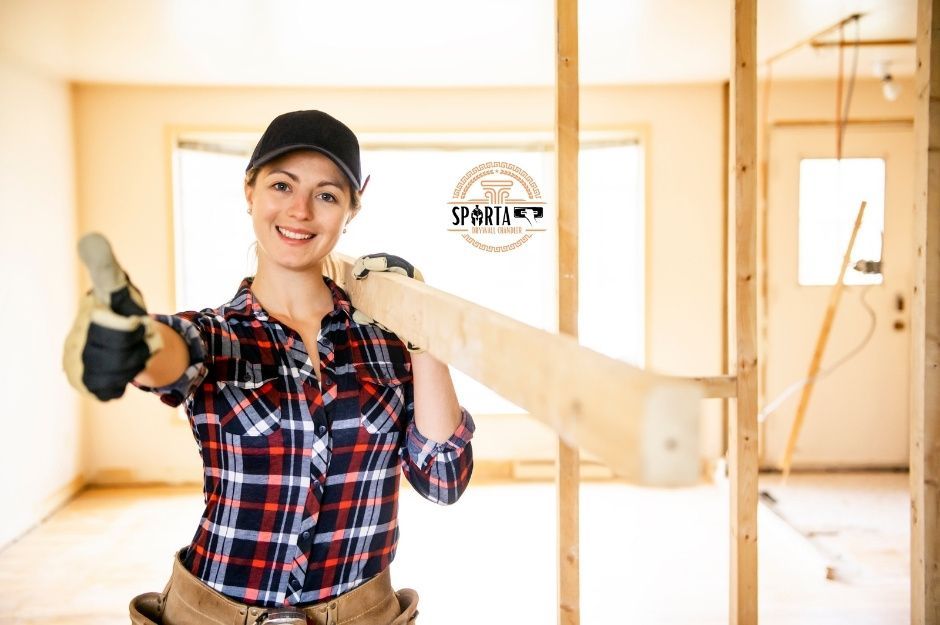 A drywall company worker in plaid shirt and baseball cap gives thumbs up while holding piece of wood at a residential construction site with exposed framing and bright natural lighting near W Farmhand Dr, and S 53rd Dr, Laveen, AZ.