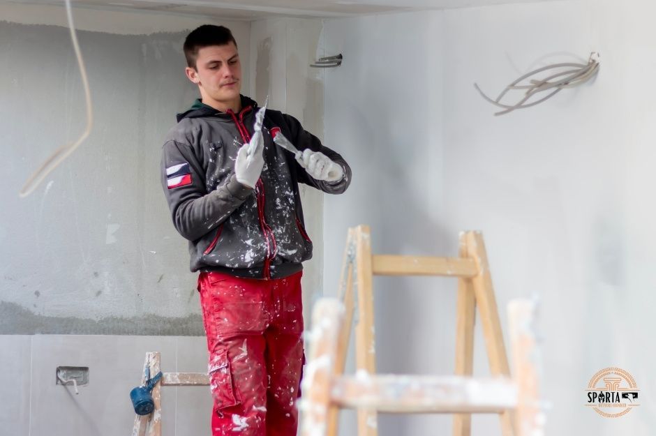 A professional drywall contractor wearing a gray hoodie and red pants uses a sanding tool on white drywall walls in a construction site. He is working on a project near Jefferson Ave, and Wicks Ave, Mayer, AZ.