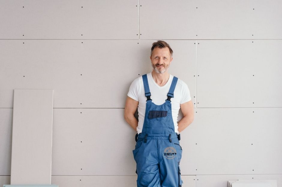 A professional drywall contractor in blue work overalls and white t-shirt stands against unfinished drywall with visible seams and screw holes. This drywall contractor is working on a project near E Pampa Ave, and S Wesley, Mesa, AZ.
